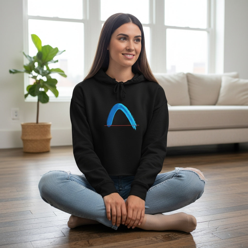 Woman sitting on the floor wearing a black crop hoodie with a turquoise arch graphic, in a bright room with a couch and plant.