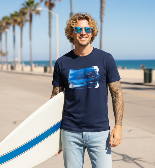 Man holding a surfboard wearing a navy t-shirt with a blue graphic design
(wave) standing on a beach.