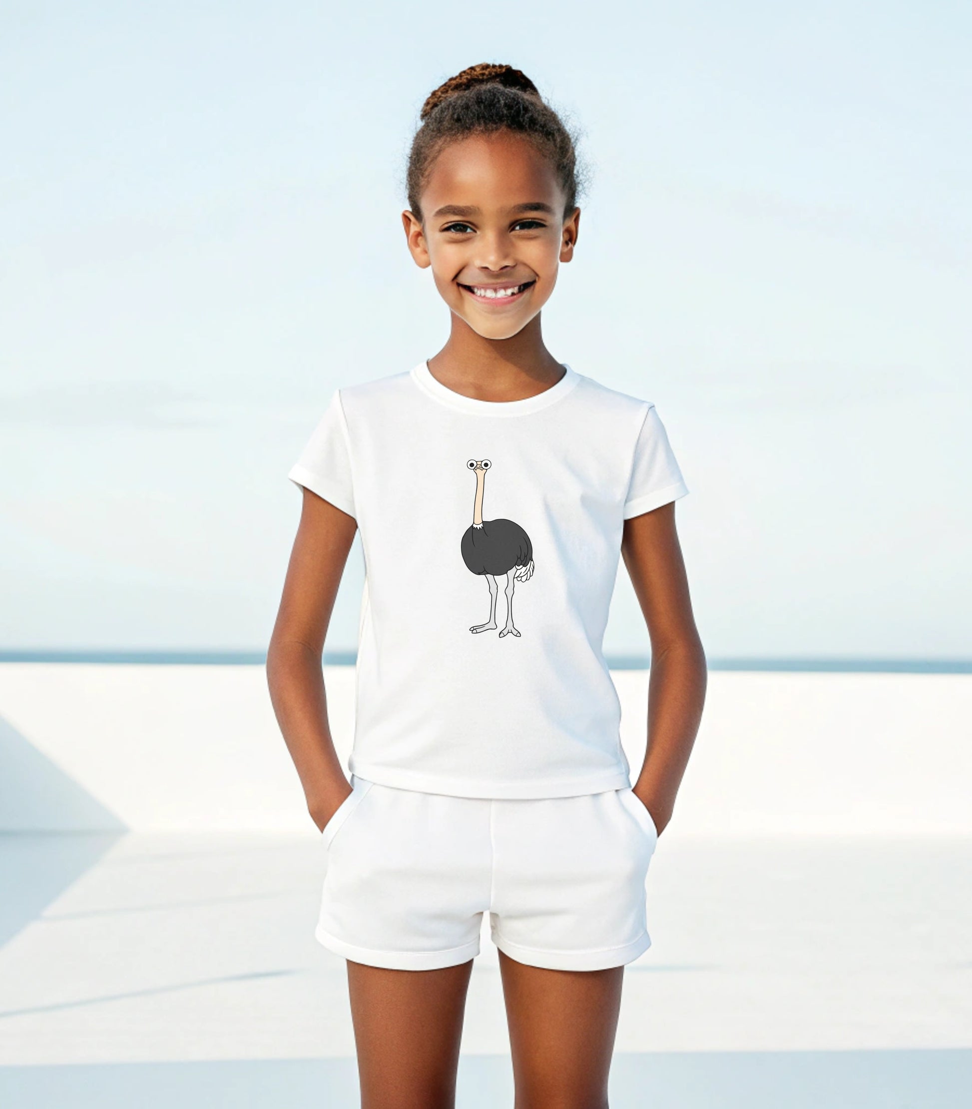 Young girl wearing a white t-shirt with an ostrich graphic and white shorts on a beach.